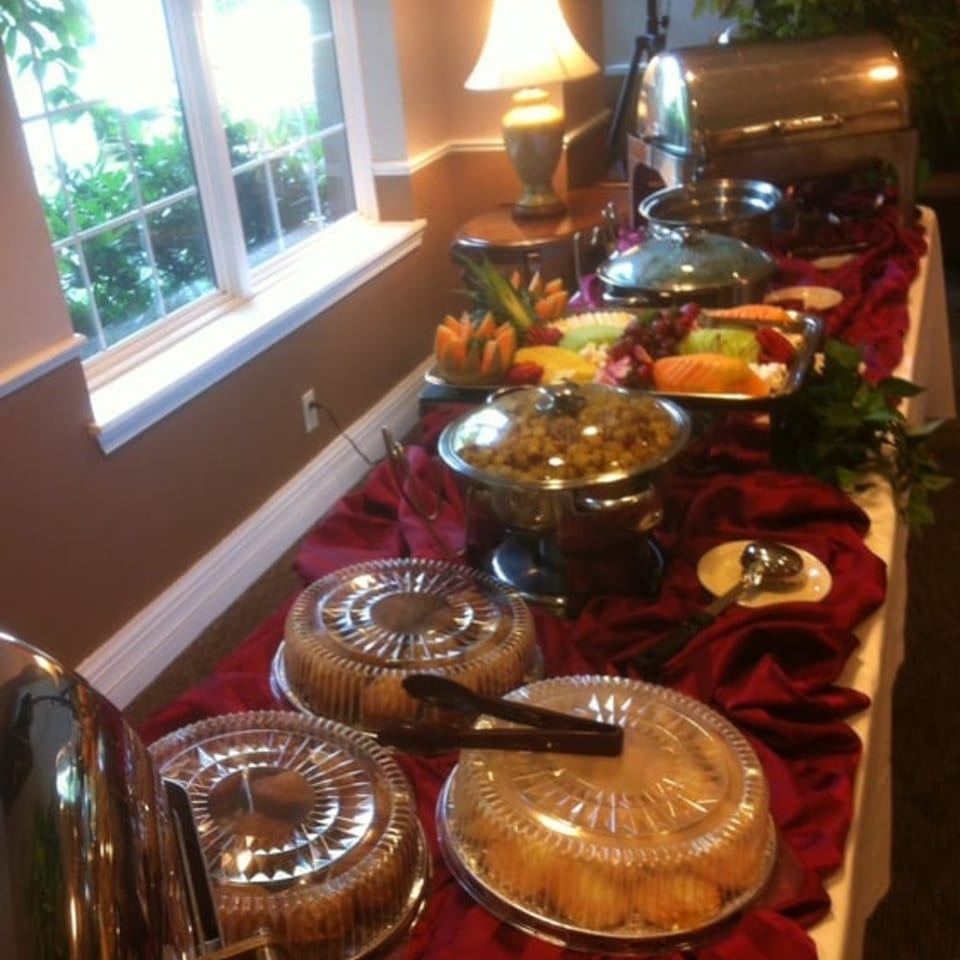 A buffet table with soul food dishes, fruit, and desserts, draped in red cloth, beside a window.