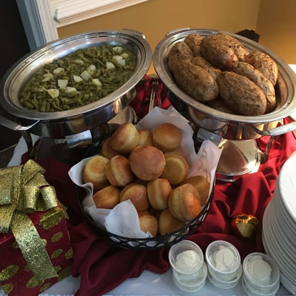 Chafing dishes of green beans and baked potatoes, with a basket of dinner rolls and butter cups.
