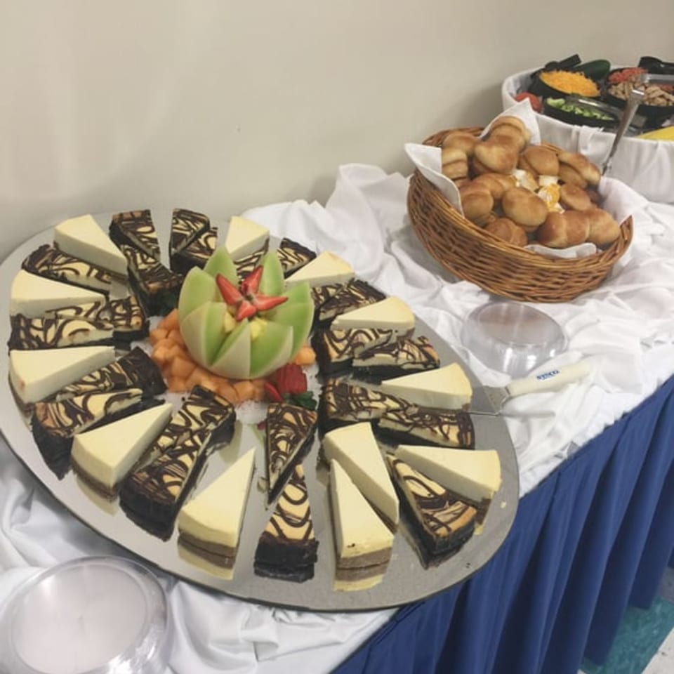 A buffet table with cheesecake slices, fruit, and a basket of dinner rolls.