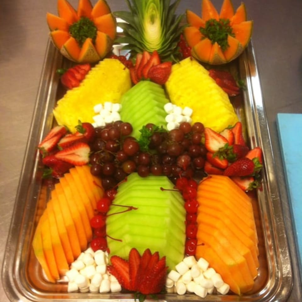 A large silver tray holds an elaborate fruit platter with carved melons, pineapple, grapes, and strawberries.